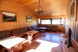 Gemütlicher Wohnraum in einem Holz-Campinghaus mit Couch, Sesseln, Tisch, Küchenzeile und großen Fenstern.