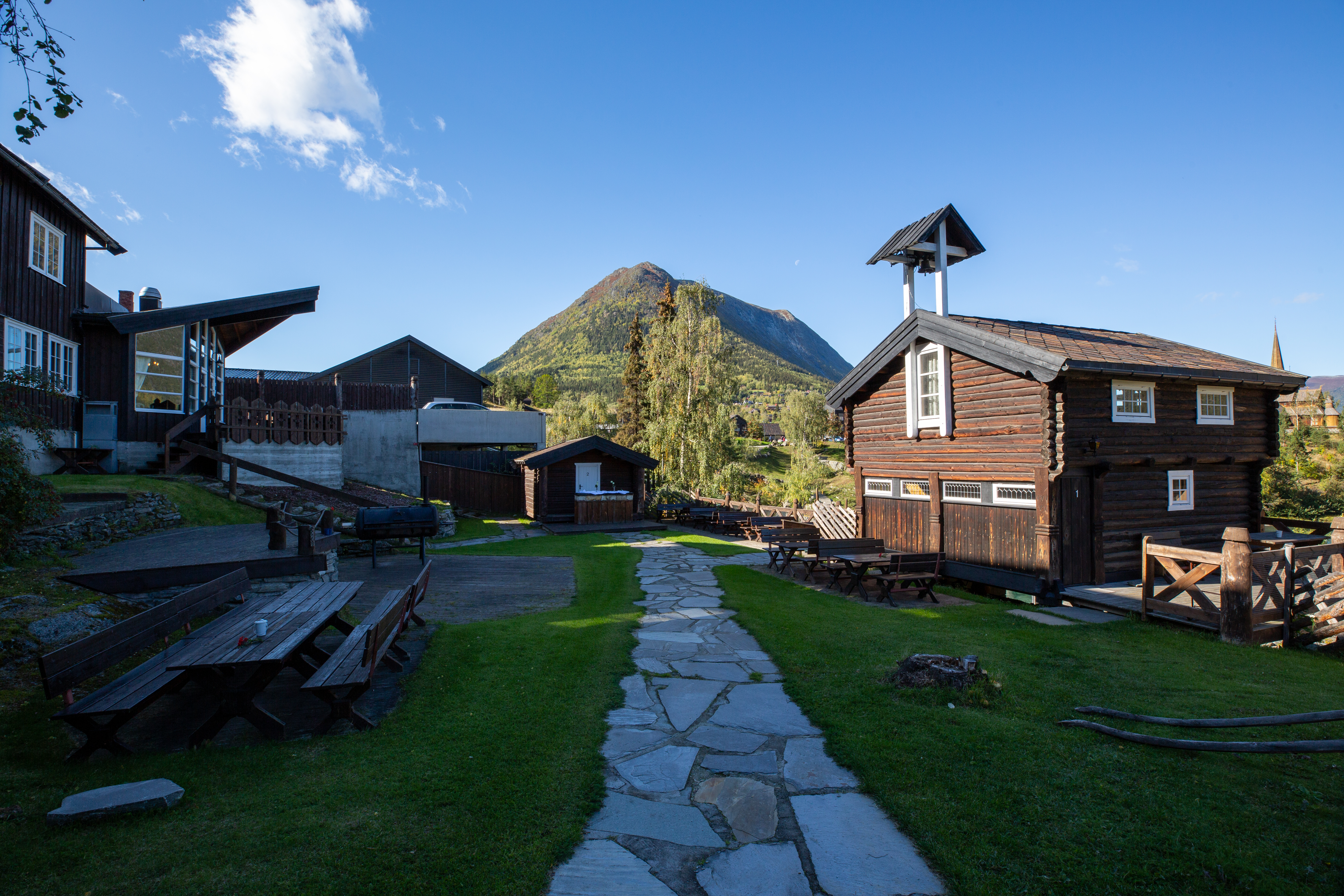 Holzhütten auf einem Campingplatz, gepflasterter Weg und Rasenfläche, im Hintergrund ein Berg und ein Holzschild.