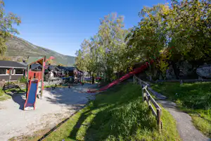 Spielplatz mit roter Rutsche am Campingplatz, Kiesweg und Holzgeländer, Bäume und mehrere Wohnwagen in der Umgebung.