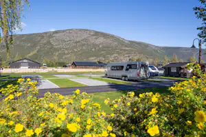 Campingplatz mit Wohnwagen und Mobilheimen, Rasenwegen und Blumenbeeten, im Hintergrund Berge und Bäume.