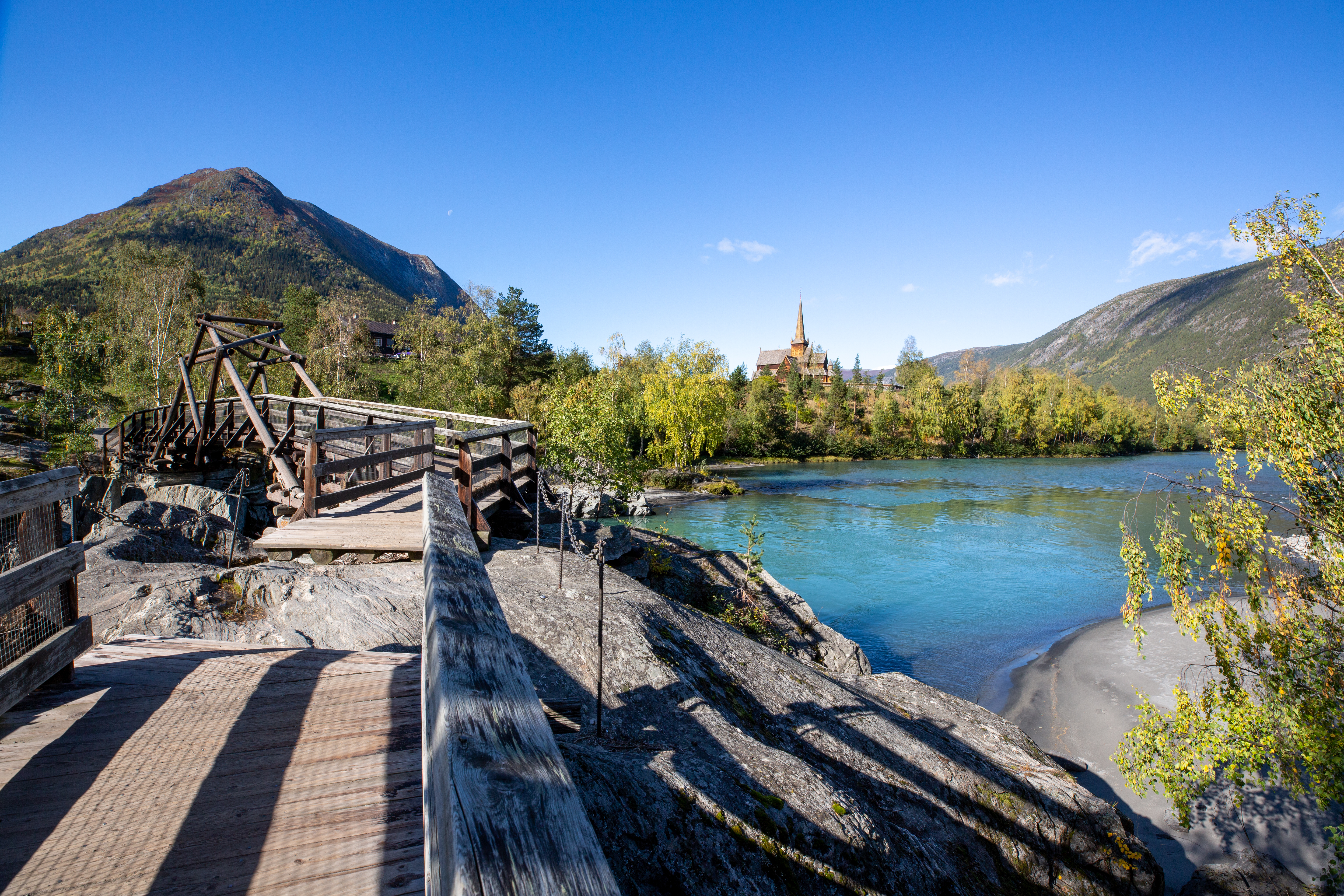 Holzsteg am Fluss mit Blick auf das Wasser, Berge und eine Kirche in der Ferne, umgeben von Bäumen.