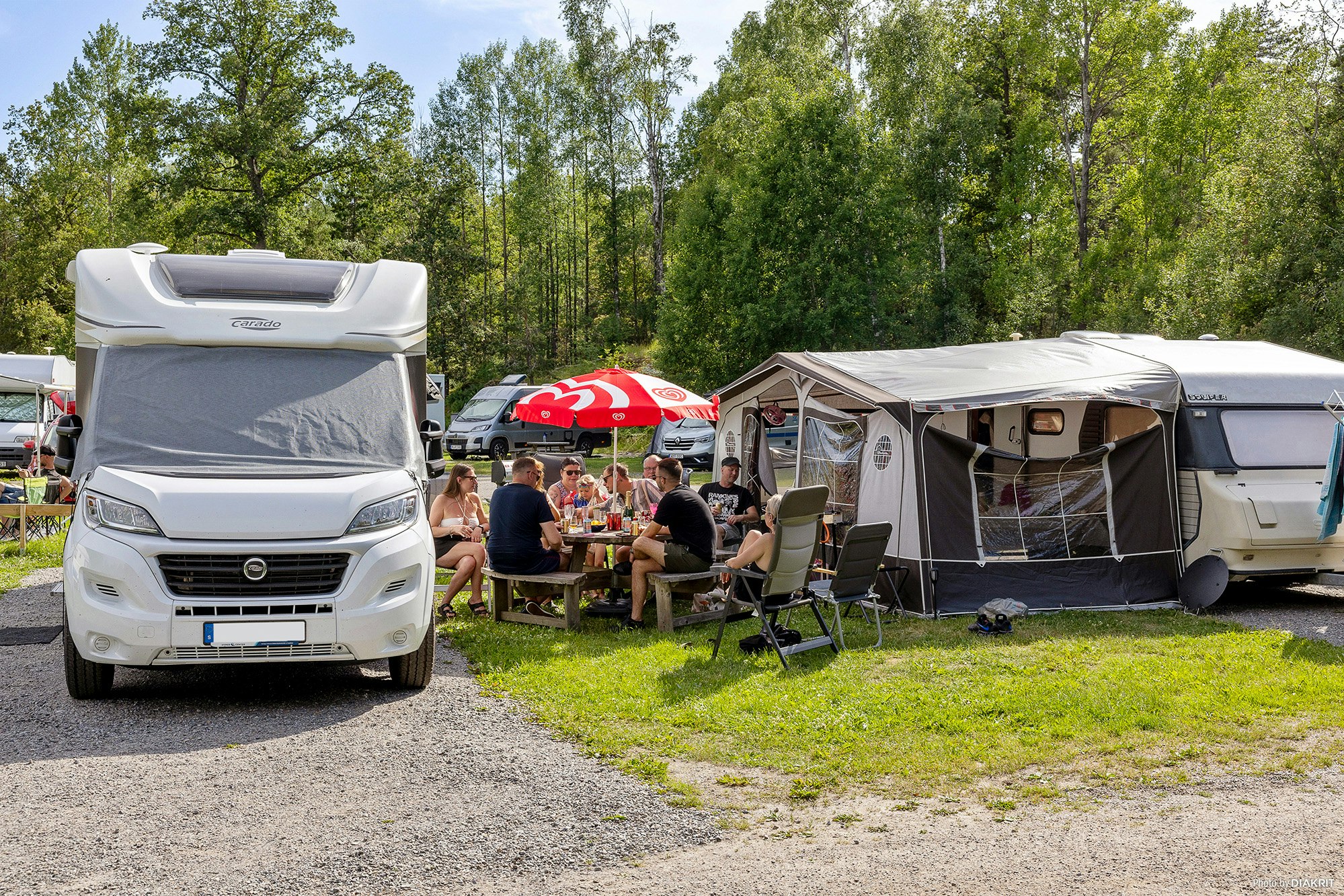 Wohnmobil mit geöffnetem Aufbau, daneben Zelte und Campingstühle; am Tisch sitzen mehrere Personen vor dem Wohnwagen.
