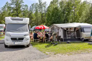 Wohnmobil mit geöffnetem Aufbau, daneben Zelte und Campingstühle; am Tisch sitzen mehrere Personen vor dem Wohnwagen.