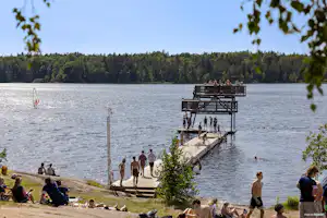 Menschen am Ufer eines Sees, im Hintergrund ein Steg mit Plattform und Holzgeländer, umgeben von Wald und Bäumen.