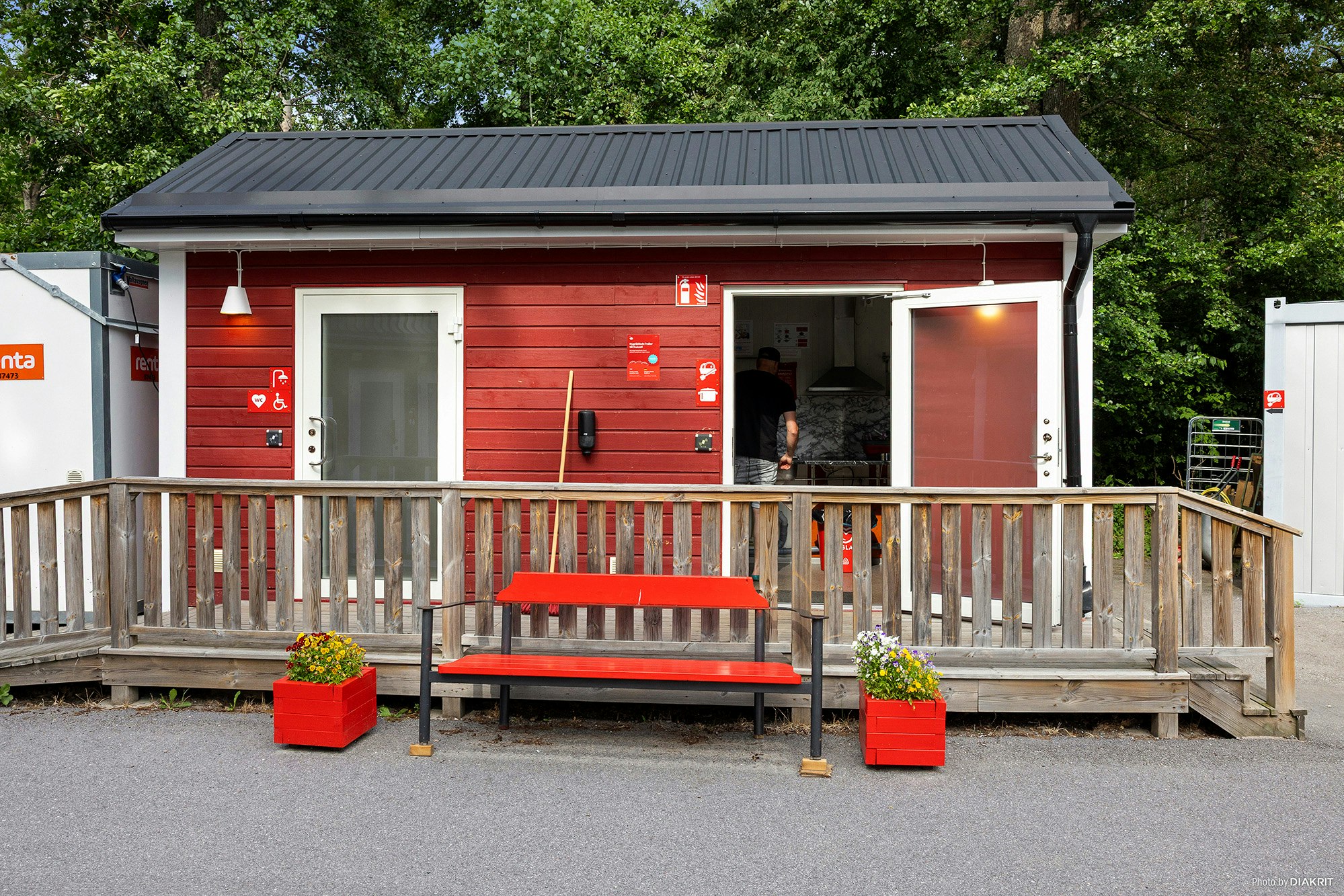 Holzhaus mit Veranda und Bank, roter Eingangstür, davor rote Sitzbank und Pflanzkübel, umgeben von Bäumen.