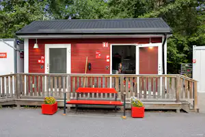 Holzhaus mit Veranda und Bank, roter Eingangstür, davor rote Sitzbank und Pflanzkübel, umgeben von Bäumen.