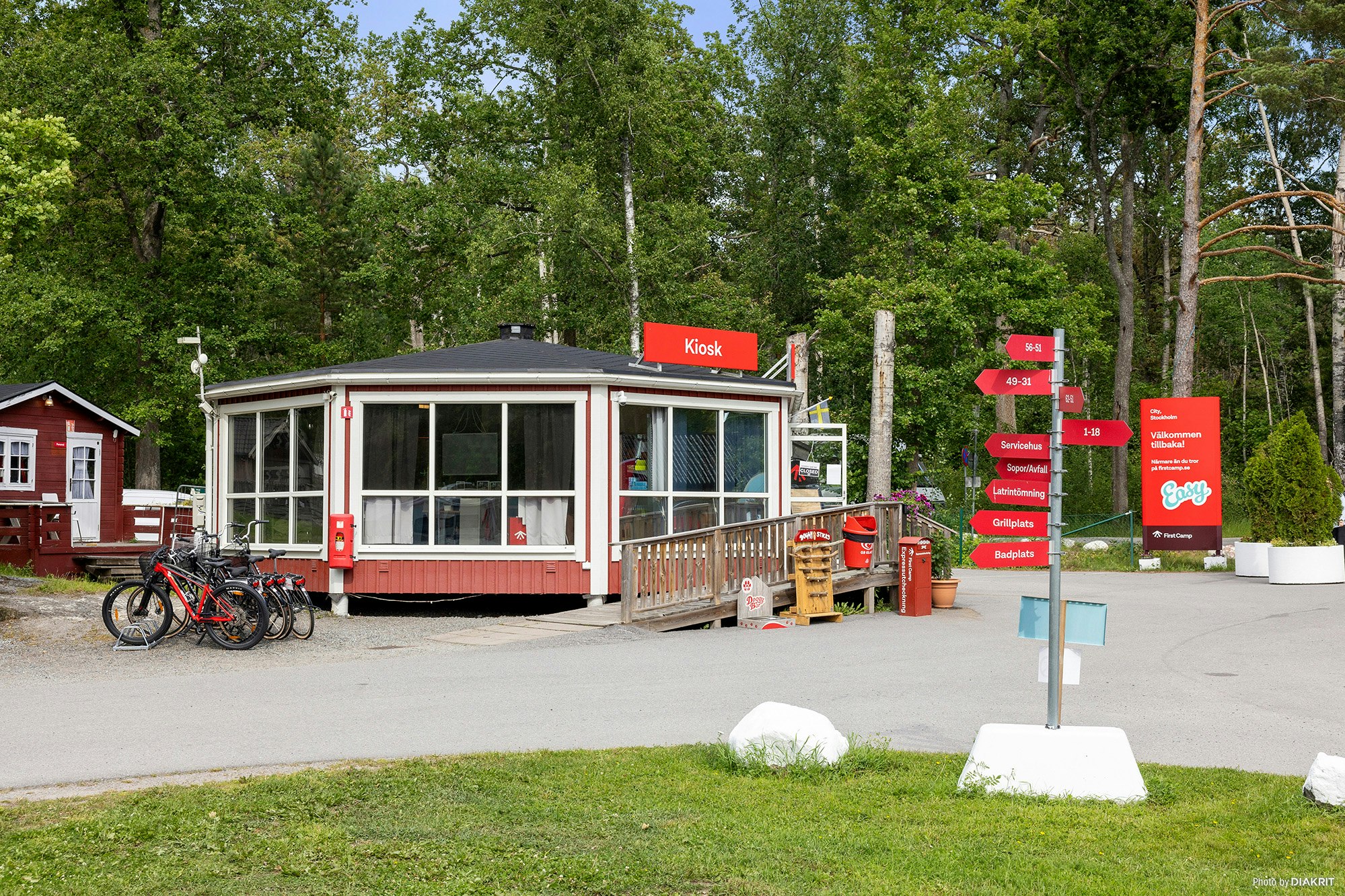 Rezeption am Campingplatz mit kleinem Gebäude, Glasveranda und roten Schildern, davor Fahrräder und Weg neben Bäumen.