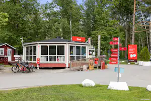 Rezeption am Campingplatz mit kleinem Gebäude, Glasveranda und roten Schildern, davor Fahrräder und Weg neben Bäumen.