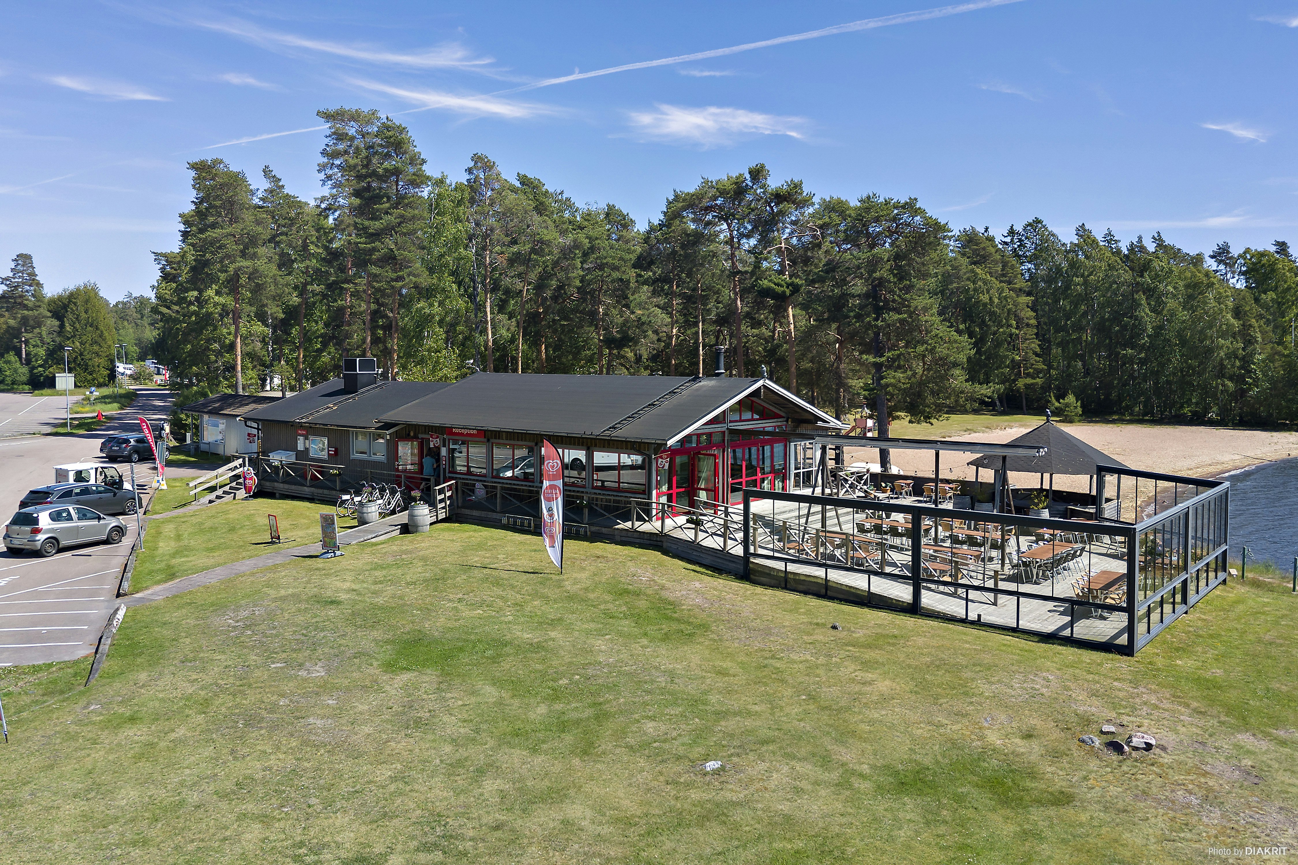 Campingplatz mit Wohnwagen und Stellplätzen, überdachter Terrasse und Grillbereich, umgeben von Bäumen und Rasenfläche.