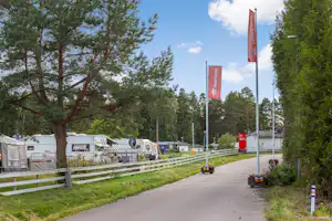 Campingplatz-Gasse mit Wohnwagen und Mobilheimen, weißen Zäunen, hohen roten Fahnen und Bäumen entlang des Weges.