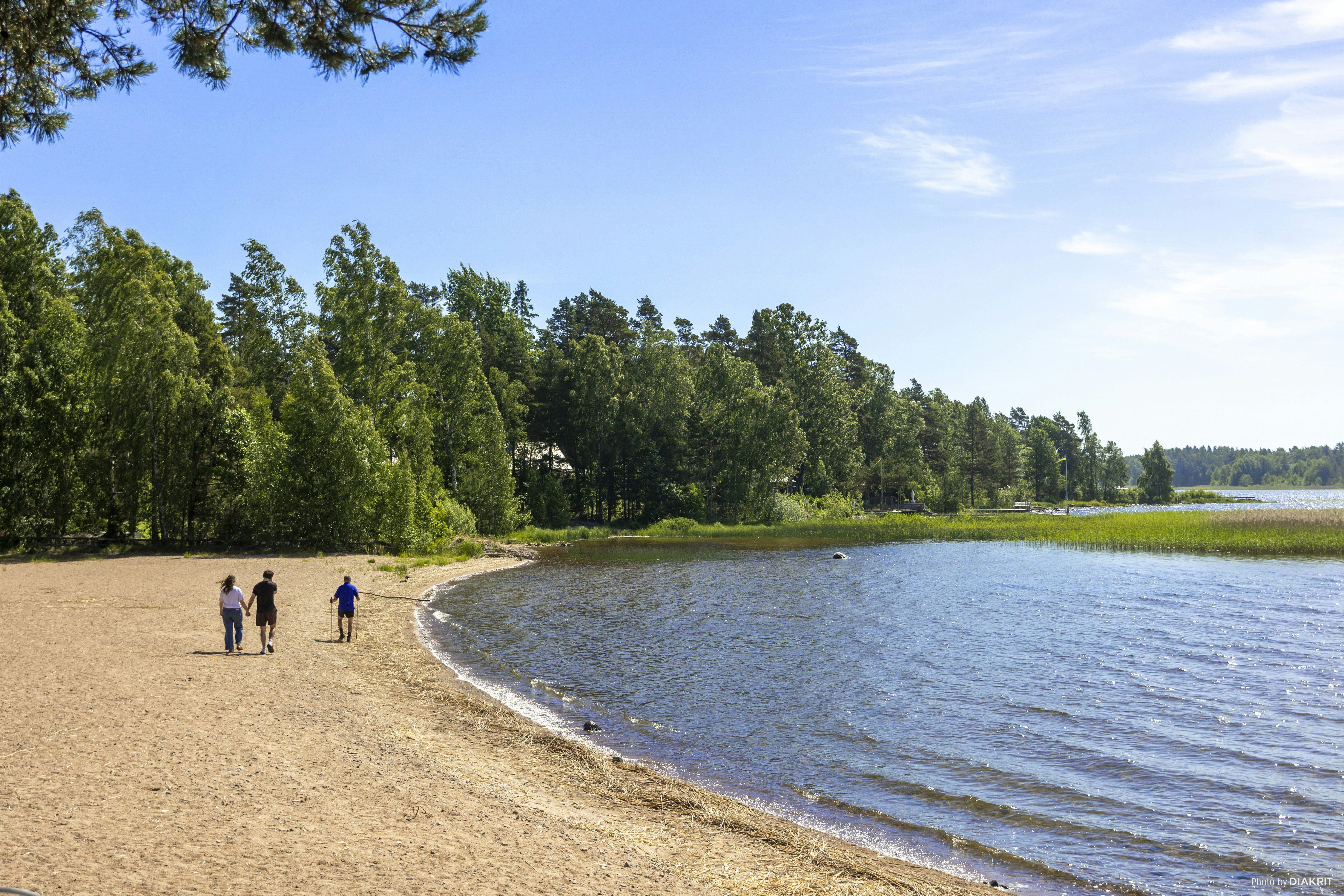 Sandstrand am See mit zwei Personen am Ufer, Bäume im Hintergrund und ruhiges Wasser.