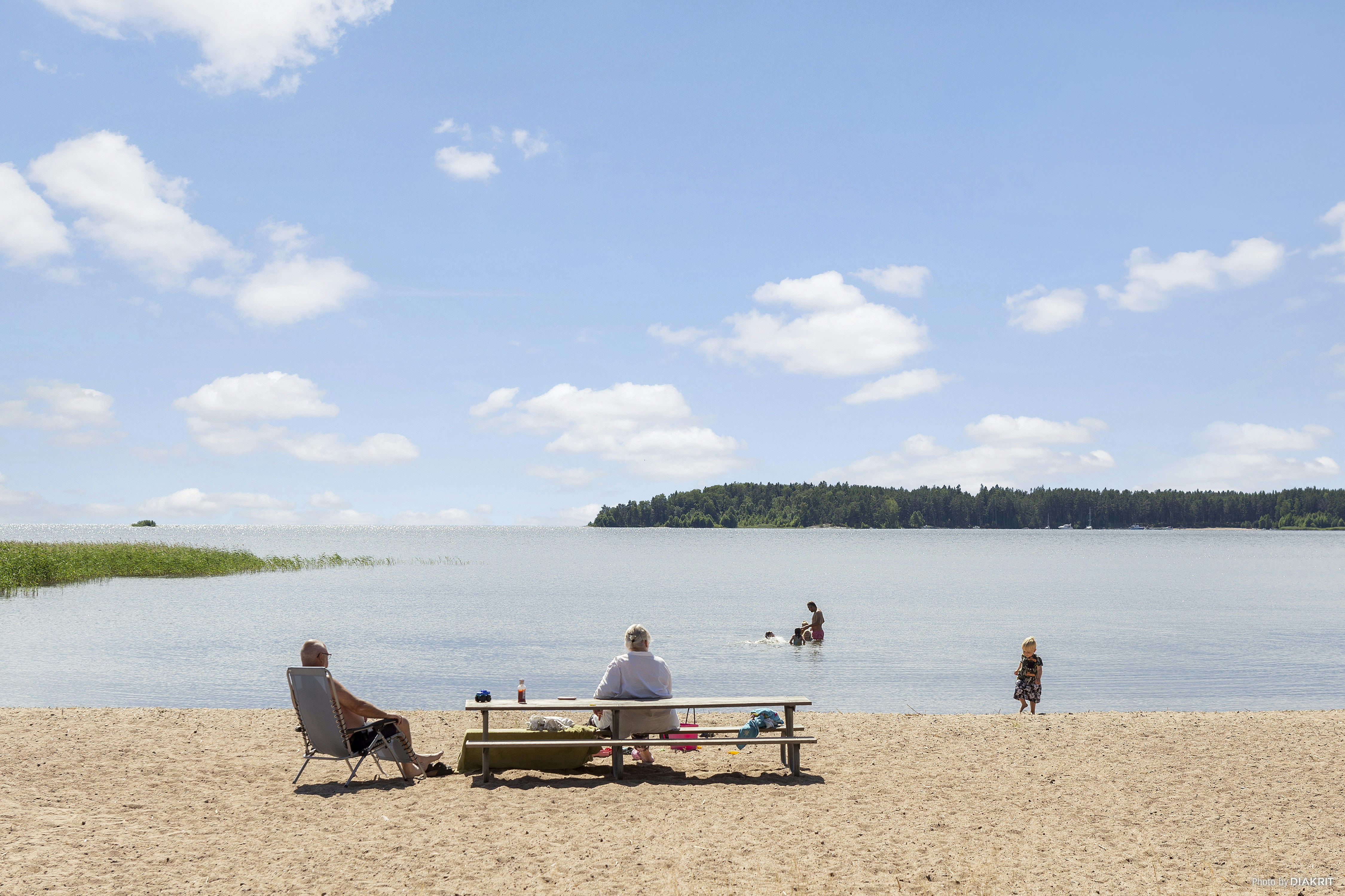 Strand am See: Liegestuhl und Tisch mit Getränken, im Hintergrund Wasser und Personen am Ufer.