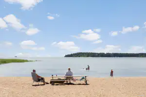 Strand am See: Liegestuhl und Tisch mit Getränken, im Hintergrund Wasser und Personen am Ufer.