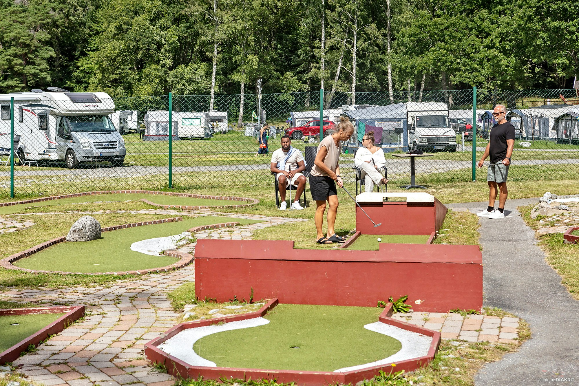 Minigolfanlage auf dem Campingplatz mit roten Bahnen, Rasenflächen und Wohnmobilen im Hintergrund, daneben ein Weg.