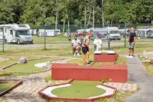 Minigolfanlage auf dem Campingplatz mit roten Bahnen, Rasenflächen und Wohnmobilen im Hintergrund, daneben ein Weg.