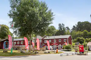 Rote Ferienhaus-Reihe auf einem Campingplatz, mit Sitzbereich, Flaggen und Bäumen im Hintergrund.