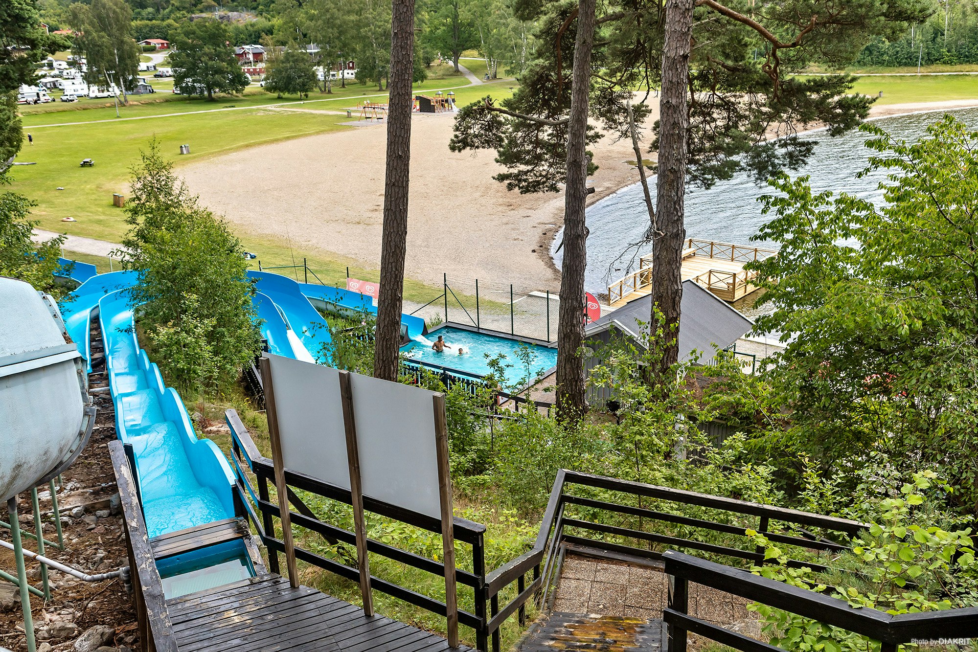 Blick vom Holzdeck auf einen Campingplatz mit Pool, blauen Rutschen und Liegen, umgeben von Bäumen und Rasenflächen.
