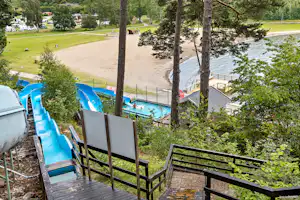 Blick vom Holzdeck auf einen Campingplatz mit Pool, blauen Rutschen und Liegen, umgeben von Bäumen und Rasenflächen.