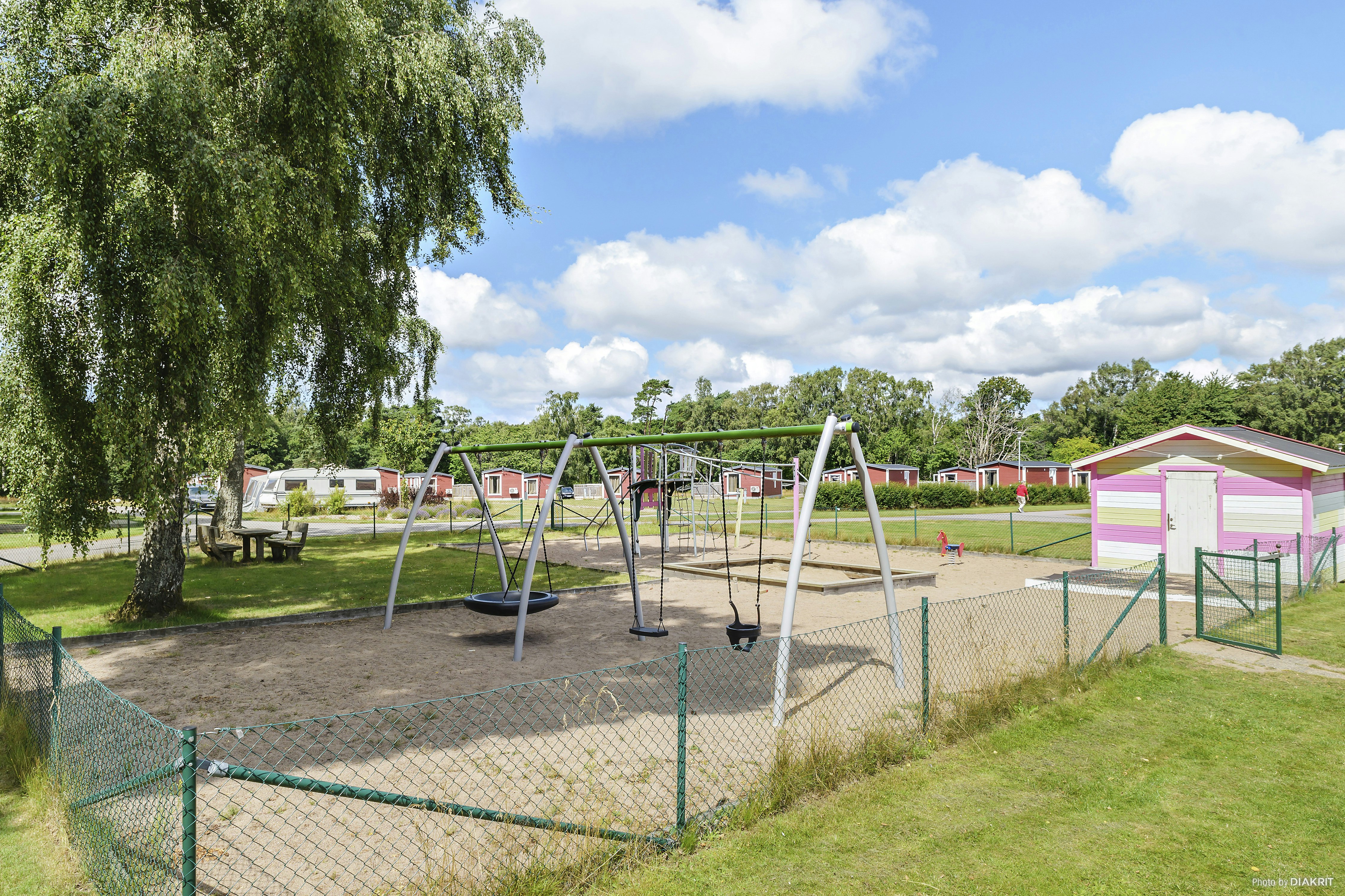 Spielplatz auf dem Campingplatz mit Trampolin, Schaukeln und eingezäuntem Bereich, Rasen und Bäumen im Hintergrund.