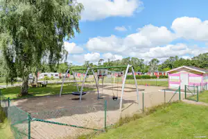 Spielplatz auf dem Campingplatz mit Trampolin, Schaukeln und eingezäuntem Bereich, Rasen und Bäumen im Hintergrund.