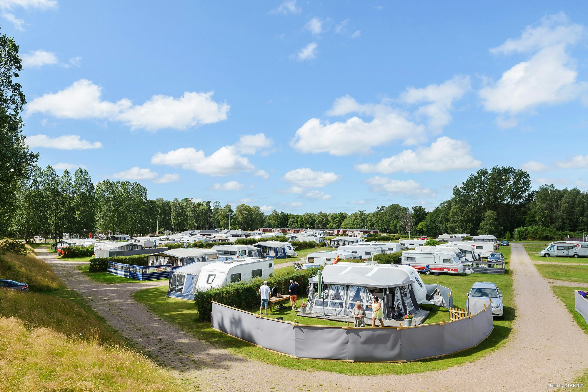 Wohnwagen und Zelte auf einem Campingplatz, umgeben von Hecken und Bäumen, mit Wegen zwischen den Stellplätzen.