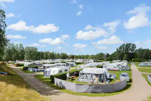 Wohnwagen und Zelte auf einem Campingplatz, umgeben von Hecken und Bäumen, mit Wegen zwischen den Stellplätzen.