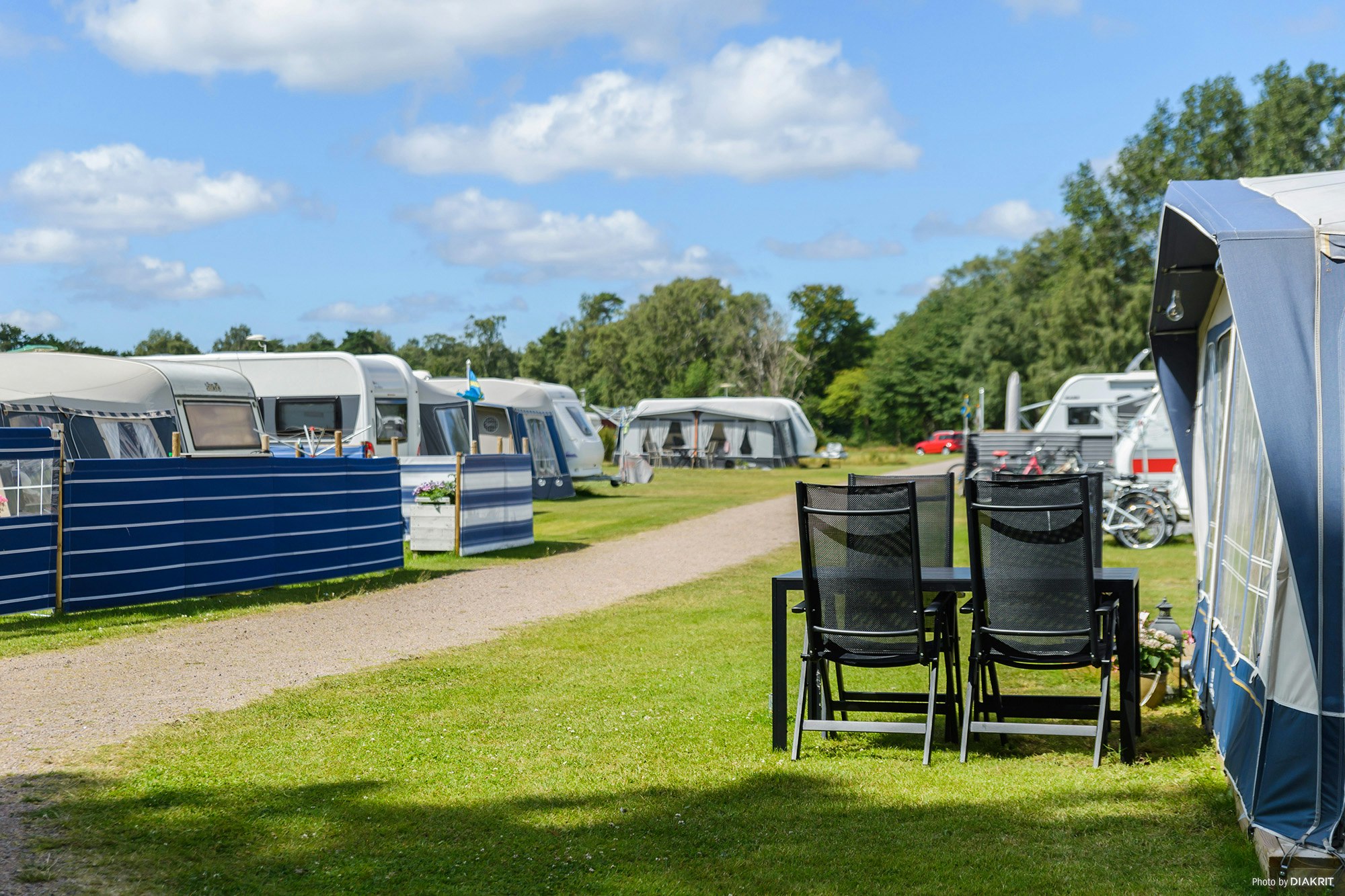 Campingplatz mit Wohnwagen und Zelten, Kiesweg und Rasenfläche; rechts zwei Campingstühle vor einem Zelt.