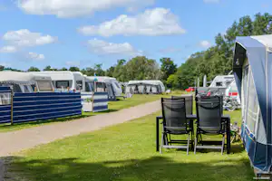 Campingplatz mit Wohnwagen und Zelten, Kiesweg und Rasenfläche; rechts zwei Campingstühle vor einem Zelt.