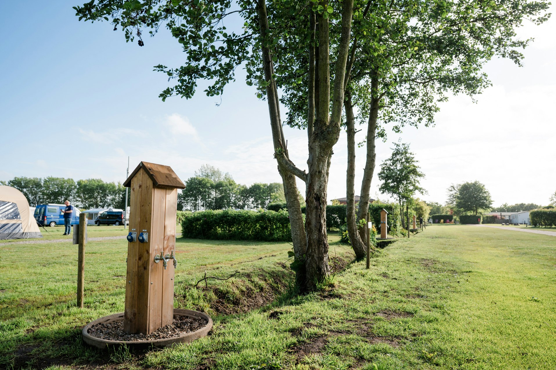 Holzschrank am Campingplatz im Grünen, neben Bäumen und Rasenfläche, im Hintergrund Zelte und ein Weg.