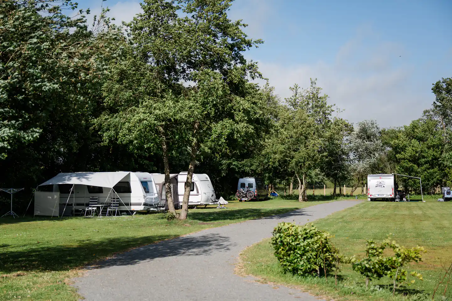 Campingplatz mit mehreren Wohnwagen und Zelten, Rasenfläche, Bäumen und einem breiten Weg zwischen den Stellplätzen.