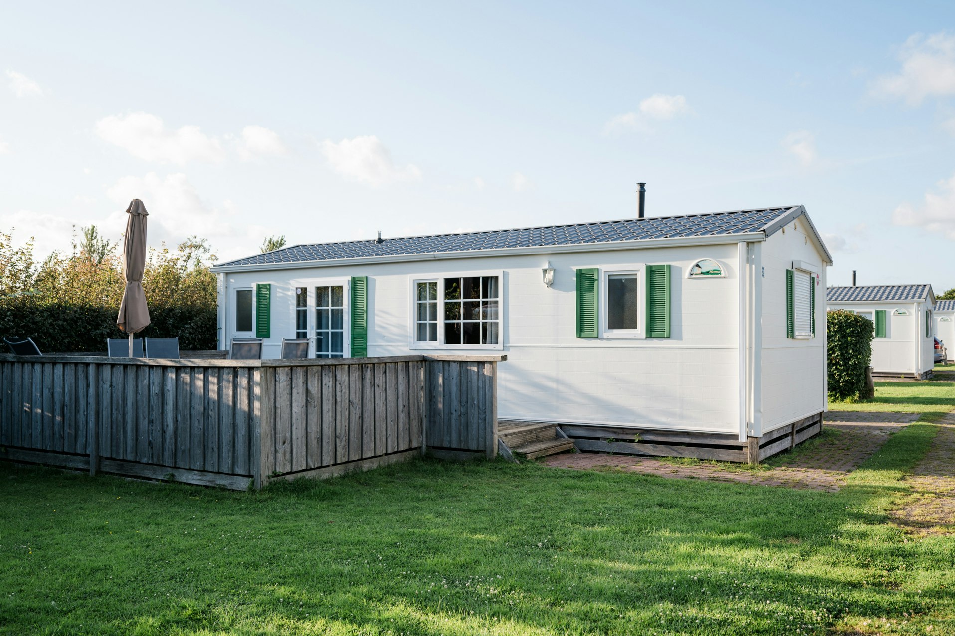 Weiße Ferienwohnung im Campingpark mit Holzsichtschutz, Rasenfläche und gepflastertem Weg, seitlich mit Fenstern und Eingangstür.