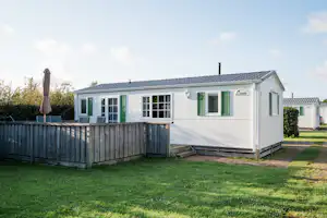 Weiße Ferienwohnung im Campingpark mit Holzsichtschutz, Rasenfläche und gepflastertem Weg, seitlich mit Fenstern und Eingangstür.