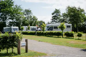 Reihe von Wohnmobilen auf einem Campingplatz, gepflasterter Weg und Hecken, im Hintergrund Bäume und ein grüner Rasen.