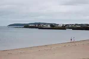 Küstenort am Meer mit Strand, Häusern am Ufer und grünen Hügeln im Hintergrund.