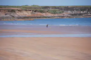 Weite Sandbucht mit einer einzelnen Person am Strand, dahinter niedrige Hügel und Felsklippen.