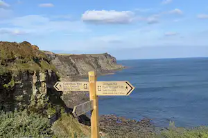 Wegweiser an den Klippen mit Blick auf das Meer und die Küste, Felsformationen und Pfad am Abhang.