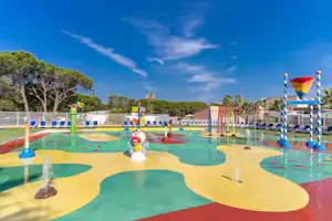Wasserspielplatz mit bunten Fontänen und Becken auf dem Campingplatz, umgeben von Bäumen und blauem Himmel.
