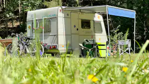 Wohnmobil auf einem Campingplatz mit Vorzelt, Campingstühlen und Fahrrädern im Vordergrund, umgeben von Bäumen.