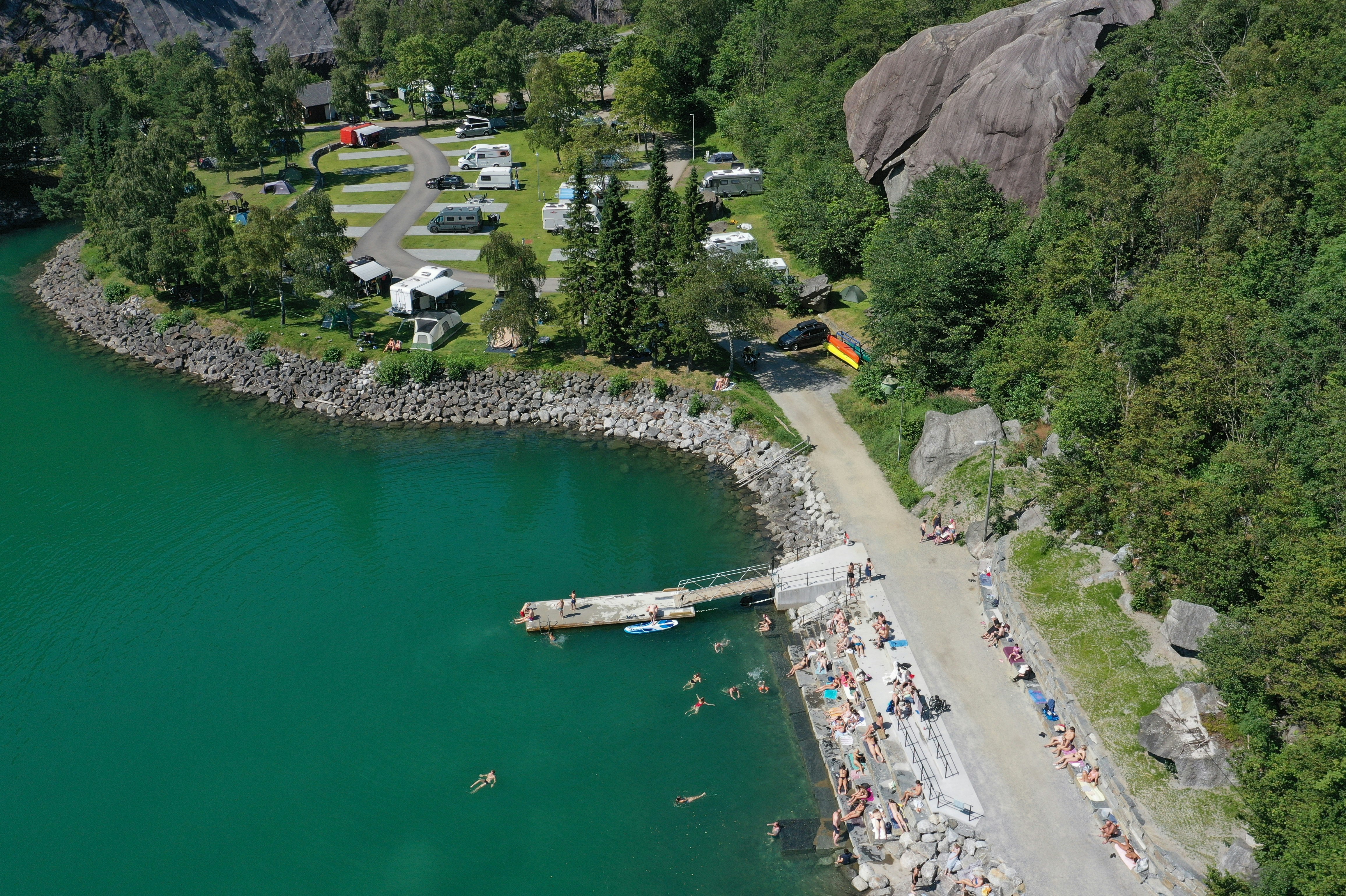 Campingplatz am See mit Steg, Booten und Uferweg, daneben Stellplätze und Wohnwagen zwischen Bäumen und Felsen.