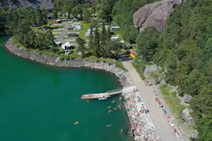Campingplatz am See mit Steg, Booten und Uferweg, daneben Stellplätze und Wohnwagen zwischen Bäumen und Felsen.