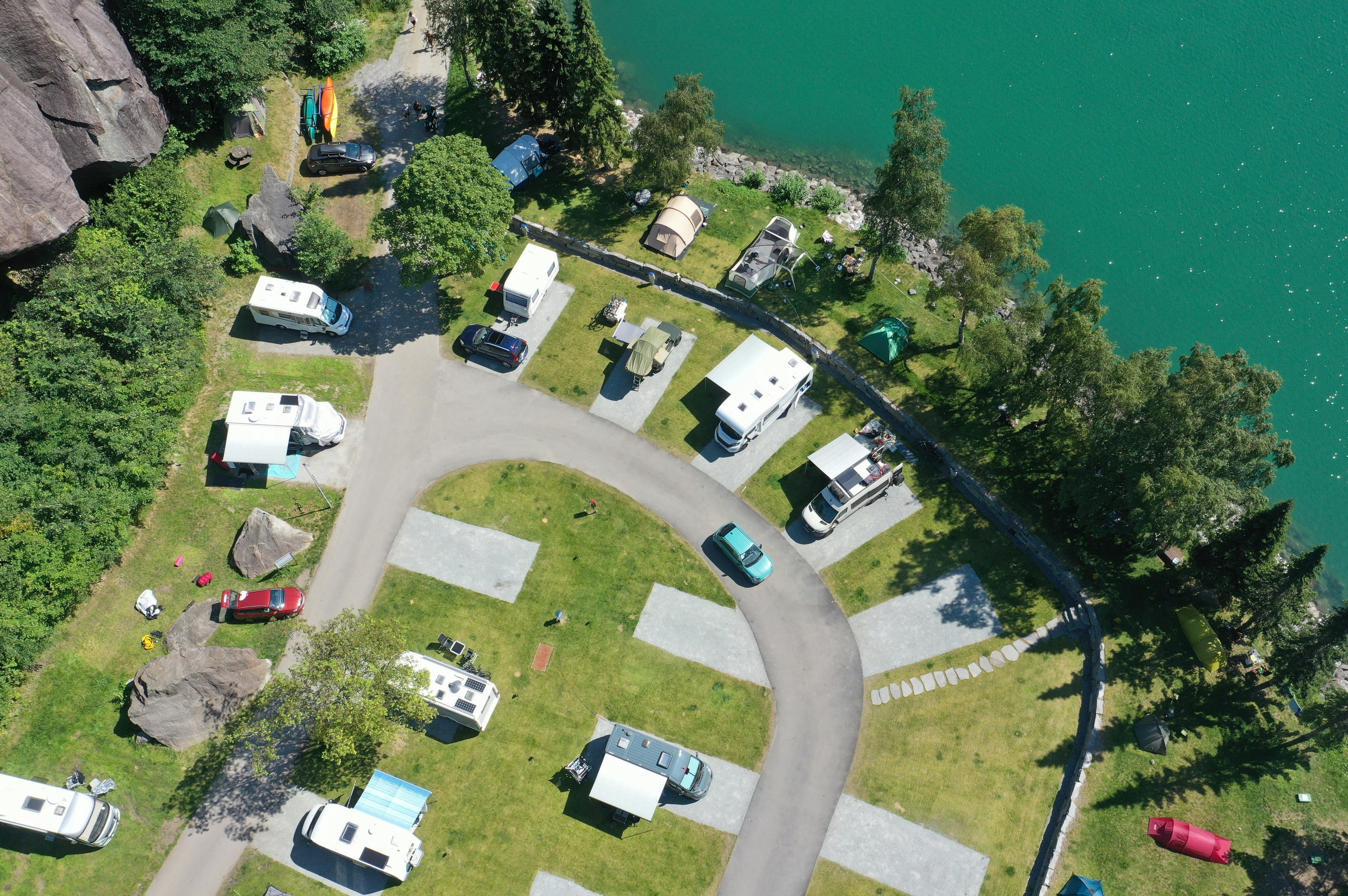 Luftaufnahme eines Campingplatzes am See mit Stellplätzen, kleinen Wohnwagen und Zelten, Wegen und Bäumen am Ufer.