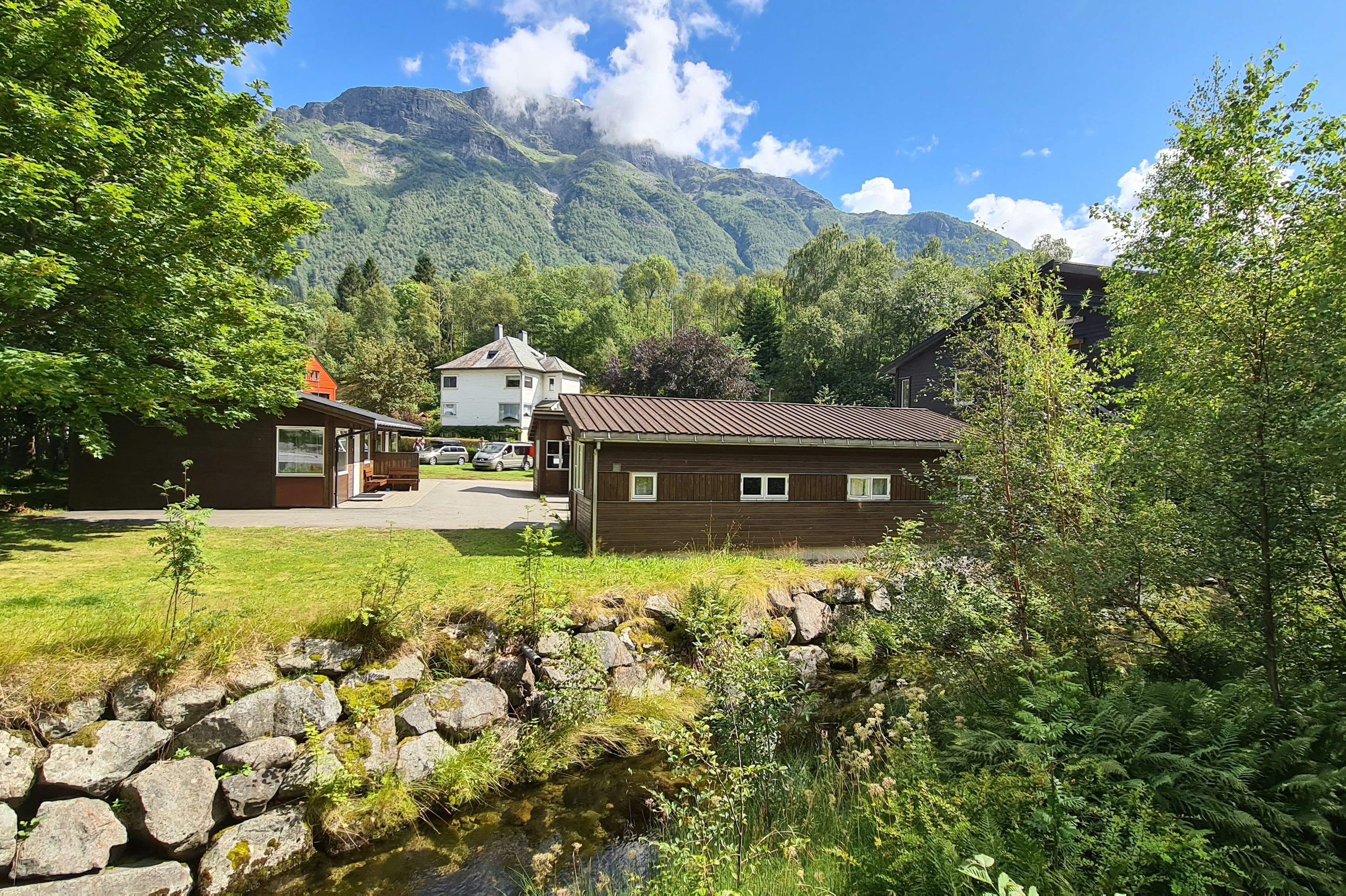 Campinganlage mit mehreren Häusern, gepflegtem Rasen und Steinmauer, umgeben von Bäumen und Bergen im Hintergrund.