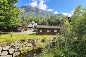 Campinganlage mit mehreren Häusern, gepflegtem Rasen und Steinmauer, umgeben von Bäumen und Bergen im Hintergrund.