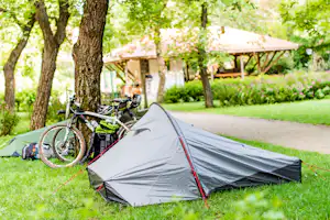 Campingzelt auf dem Rasen, davor ein Fahrrad, im Hintergrund Bäume und ein überdachter Bereich am Stellplatz.