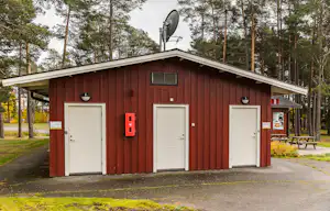 Rotes Campinggebäude mit drei weißen Türen, Alarmmelder und Lautsprecher an der Fassade, umgeben von Bäumen und Wegen.