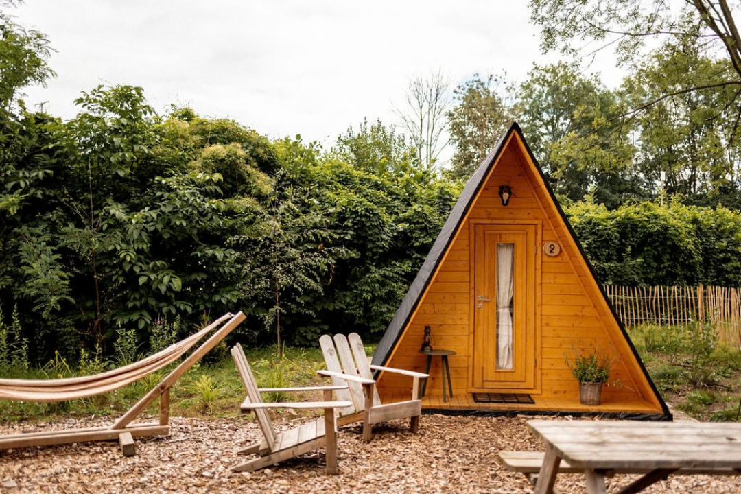 Dreieckiges Holz-Zelt mit Eingangstür, davor zwei Campingstühle und Tisch auf Kies, umgeben von Bäumen und Hecken.
