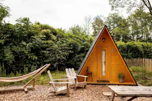 Dreieckiges Holz-Zelt mit Eingangstür, davor zwei Campingstühle und Tisch auf Kies, umgeben von Bäumen und Hecken.