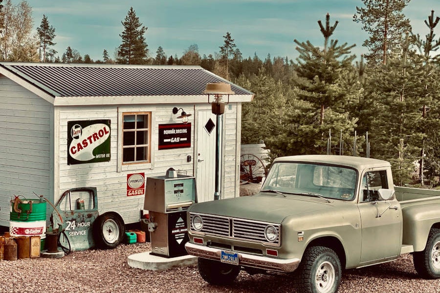 Alter Campingwagen mit Aufklebern und geöffneter Tür, daneben ein weißer Oldtimer auf dem Stellplatz, umgeben von Bäumen.