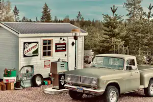 Alter Campingwagen mit Aufklebern und geöffneter Tür, daneben ein weißer Oldtimer auf dem Stellplatz, umgeben von Bäumen.