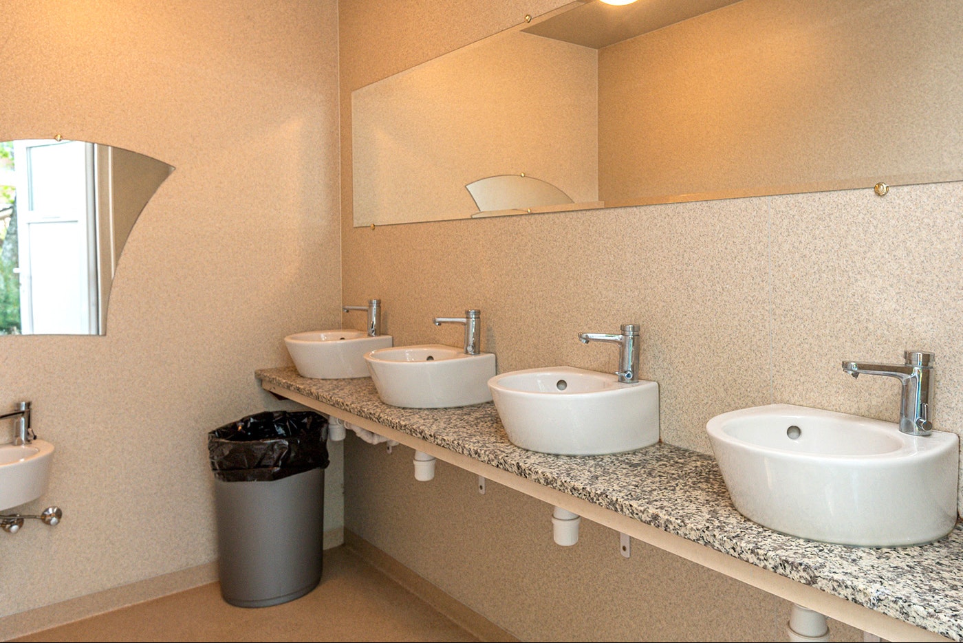Mehrere Waschbecken in einem modernen Sanitärraum, mit Steinwaschtisch, Armaturen und Abfalleimer an der Wand.